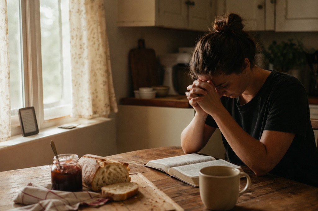 Interior of a warm Christian family kitchen at dawn: open Bible on a wooden table, coffee mug, a small e-ink Lamp on the windowsill, bread and jam, a figure at quiet prayer.