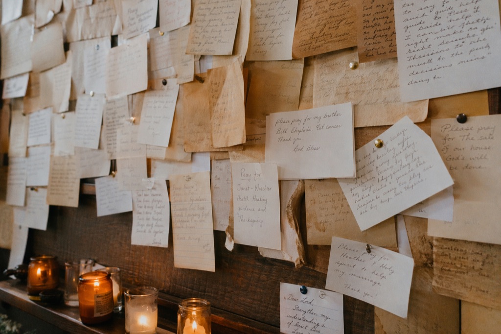 A wall of handwritten prayer cards in varied papers and handwriting, votive candles glowing on a wooden shelf below.