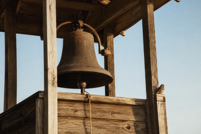 A bronze bell hanging in a simple wooden tower in morning light, a rope dangling, a bird perched on the tower's edge.