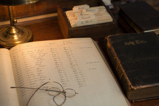 A handwritten church ledger open on a wooden desk, columns of names and dates in fountain-pen ink, brass desk lamp, leather-bound family Bible.