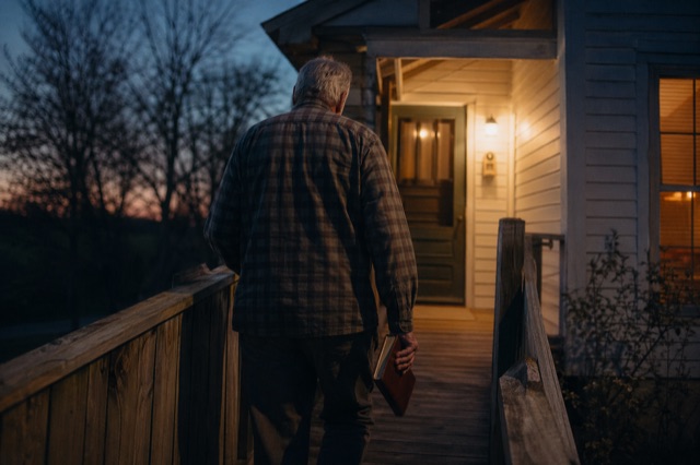 An older man in a worn plaid shirt walking slowly up a wooden walkway at twilight, a leather Bible in one hand.