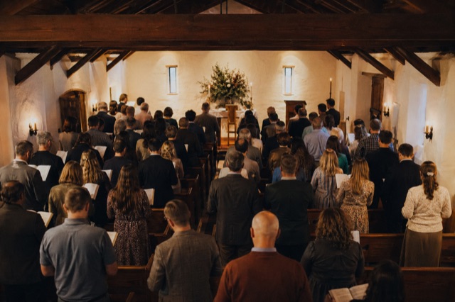 A chapel full of people standing and singing, backs to camera, warm light, wood-beamed ceiling.