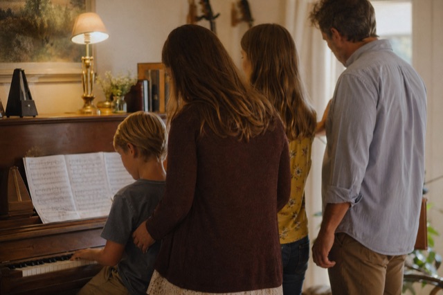 A small family of four around an upright piano at home, child's hands on the keys, warm domestic light.