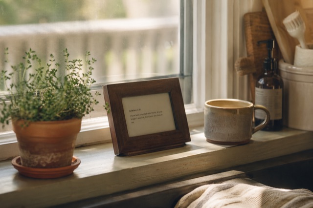A small e-ink display in a hand-finished walnut frame on a kitchen windowsill, morning light, coffee cup nearby.
