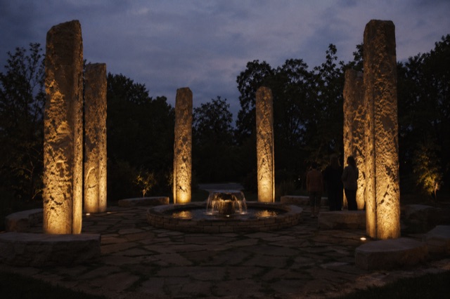 Seven tall slender stone pillars arranged in a circle around a small stone fountain at twilight, each pillar lit softly from within.