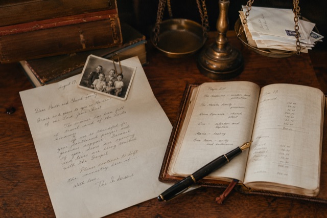 Close-up of a writing desk: a handwritten missionary letter, family photograph, leather prayer journal, fountain pen, brass scale.