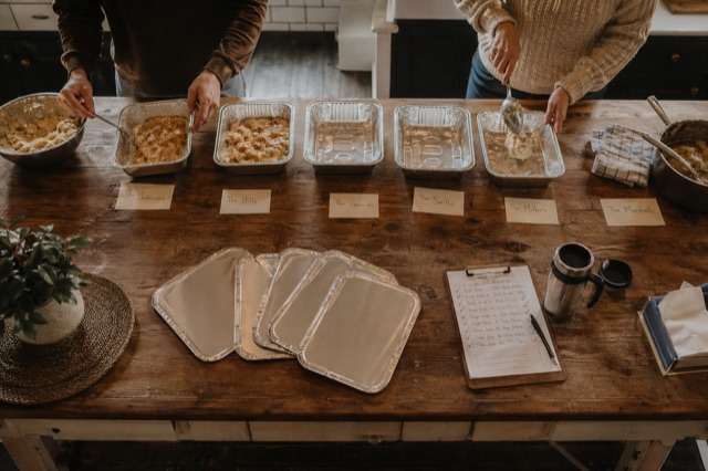 Overhead view of a community kitchen: meal-train deliveries being assembled — aluminum trays with handwritten name labels, clipboard with addresses, hands packing food.