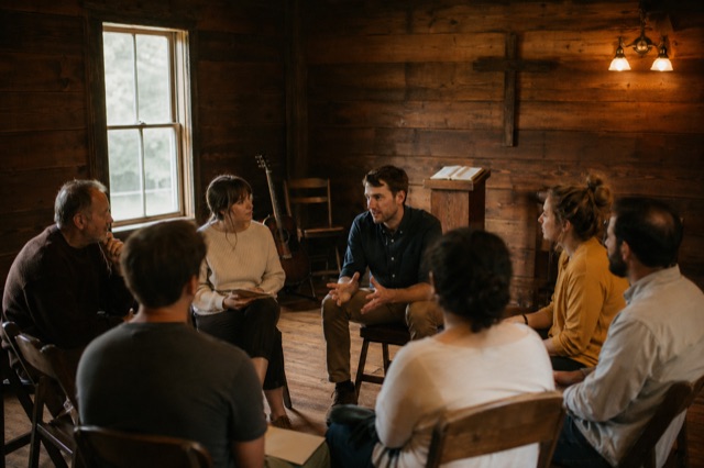 A small chapel room with warm wood walls, chairs in a rough circle, plain wooden cross, lectern with open Bible, acoustic guitar.